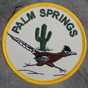 May include: Embroidered patch with a cream-colored background, featuring a roadrunner and a green cactus. The words "PALM SPRINGS" are arched above the design in green lettering. The patch has a yellow border.