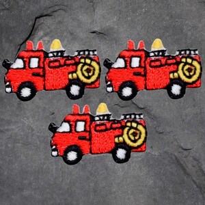 Könnte beinhalten: Drei gestickte Aufnäher von roten Feuerwehrautos mit schwarzen Rädern, weißen Scheinwerfern und gelben Akzenten. Die Aufnäher befinden sich auf einem grauen, strukturierten Hintergrund.