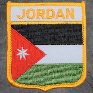 May include: Embroidered patch featuring the flag of Jordan. The patch has a white top with the word "JORDAN" in gold lettering. Below is a black stripe, a white stripe, and a green stripe. A red triangle with a white star is on the left.