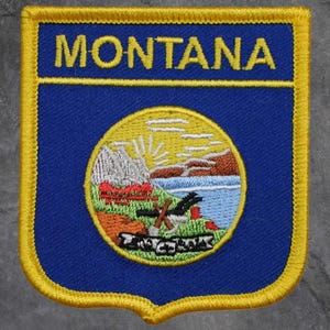 May include: Embroidered patch featuring the word "MONTANA" in yellow on a blue background. The patch has a shield shape with a yellow border. The center design includes a landscape with mountains, water, and a rising sun.