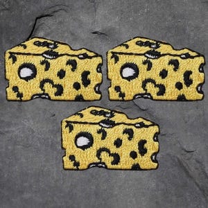 May include: Three embroidered cheese wedge patches. Each patch is yellow with black spots and white accents, resembling Swiss cheese. The patches are arranged on a gray, textured surface.