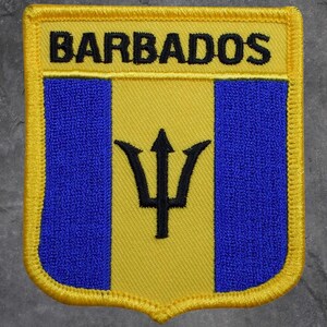 Könnte beinhalten: Gestickter Aufnäher mit der Flagge von Barbados. Das Design umfasst einen gelben oberen Bereich mit dem Wort "BARBADOS" in Schwarz, einen zentralen blauen Streifen und einen gelben unteren Bereich mit einem schwarzen Dreizack-Symbol.