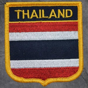 May include: Embroidered patch featuring the flag of Thailand. The patch has a shield shape with a yellow border. The flag design includes horizontal stripes of red, white, and blue, with the word "THAILAND" in yellow at the top.