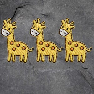 May include: Three embroidered giraffe patches. Each patch features a yellow giraffe with orange spots, a white muzzle, and a black outline. The patches are arranged in a row against a dark gray background.