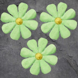 May include: Three light green daisy-shaped appliques with yellow centers are arranged on a dark gray, textured surface. The appliques have a stitched appearance, with each petal clearly defined. The overall design is simple and floral.