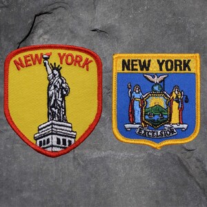 May include: Two embroidered patches featuring New York imagery. One patch shows the Statue of Liberty with "NEW YORK" in red lettering. The other patch displays the New York state seal with "NEW YORK" above and "EXCELSIOR" below.