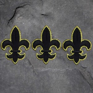 May include: Three black fleur-de-lis patches with gold embroidered outlines. The patches are arranged in a row against a textured, gray background. Each patch features a stylized, symmetrical design.
