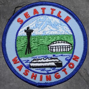 May include: Embroidered patch featuring Seattle, Washington landmarks. The design includes a mountain, Space Needle, ferry, and stadium, with "SEATTLE WASHINGTON" in a circular border. The patch is blue, red, white, green, and black.