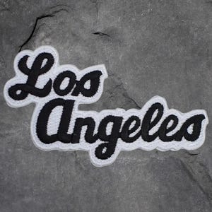 May include: A white embroidered patch with the words "Los Angeles" in a black, cursive font. The patch has a black border and is set against a gray, textured background.