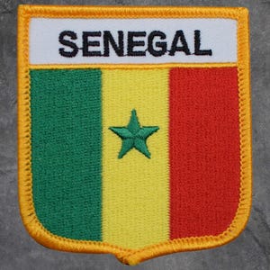 May include: A rectangular patch featuring the flag of Senegal. The patch has a white banner at the top with the word "SENEGAL" in black letters. The flag design includes green, yellow, and red vertical stripes, with a green star in the yellow stripe. The patch is bordered with a gold trim.
