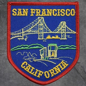 May include: Embroidered patch featuring the Golden Gate Bridge and the text "SAN FRANCISCO CALIFORNIA" in yellow lettering. The patch has a blue background with a red border, showcasing a shield shape. The design includes stylized illustrations of the bridge and other landmarks.