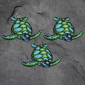 Könnte beinhalten: Drei gestickte Meeresschildkröten-Patches. Jede Schildkröte hat einen grün-blauen gemusterten Panzer, grüne Flossen und ein detailliertes Gesicht mit roten Akzenten. Die Patches befinden sich auf einer dunkelgrauen, strukturierten Oberfläche.