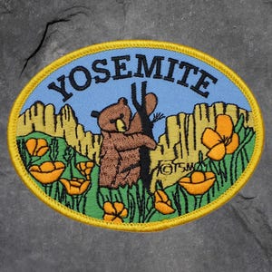 May include: Oval patch with a blue background and the word "YOSEMITE" in black. A brown bear clings to a tree, surrounded by yellow flowers and green grass. The patch has a yellow border.