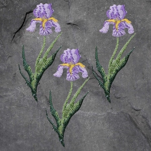 Könnte beinhalten: Drei gestickte Irisblüten mit lila Blütenblättern und gelben Akzenten. Die Blumen haben grüne Stiele und Blätter, vor einem grauen Hintergrund. Die Stickdetails sind sichtbar und zeigen die Textur und das Design des Blumenmusters.