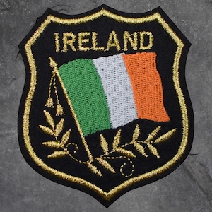 May include: Embroidered patch featuring the word "IRELAND" in gold above the Irish flag. The flag is green, white, and orange. The patch has a black background with a gold border and decorative gold leaves.