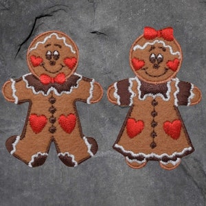 以下が含まれることがあります： 赤いハートが胸に描かれた、男性と女性のジンジャーブレッドマン2人。茶色のアイシングで装飾され、どちらも笑顔で、縁取りは白いアイシングで仕上げられています。