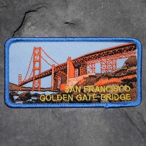 Könnte beinhalten: Gestickter Aufnäher mit der Golden Gate Bridge in San Francisco. Der Aufnäher hat einen blauen Rand und zeigt die Brücke in Orange und Braun vor einem hellblauen Himmel. Der Text "SAN FRANCISCO GOLDEN GATE BRIDGE" ist goldfarben gestickt.