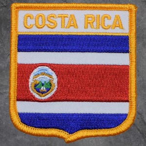 May include: Embroidered patch featuring the Costa Rican flag. The patch has a white background with the words "COSTA RICA" in gold. The flag design includes blue, white, and red stripes, and a small emblem. The patch is bordered with gold.
