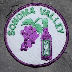 May include: Embroidered patch featuring the words "Sonoma Valley" in green, surrounding a white circle. Inside the circle are a purple bunch of grapes, a green leaf, and a purple wine bottle with a green label. The patch has a purple border.