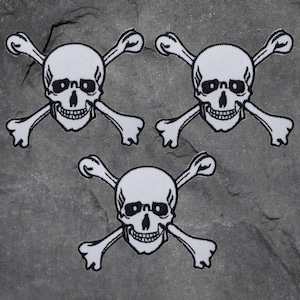 Könnte beinhalten: Drei weiße Totenkopf- und gekreuzte Knochen-Patches mit schwarzen Umrissen. Jeder Patch zeigt einen detaillierten Schädel mit Augenhöhlen, einer Nasenhöhle und einem grinsenden Mund, der von zwei Knochen gekreuzt wird. Auf grauem Hintergrund.