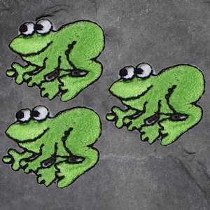 May include: Three embroidered green frog patches with white and black detailing. Each frog has large, expressive eyes and a smiling mouth. These patches are suitable for clothing or craft projects.