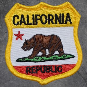 Peut inclure: Écusson brodé en forme de bouclier, avec le mot "CALIFORNIA" en noir sur fond jaune. Il comprend un ours brun, une étoile rouge et le mot "REPUBLIC" en rouge, représentant l'État.