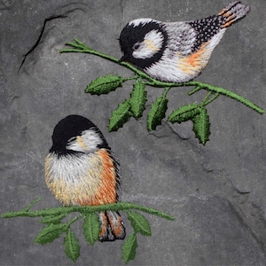 May include: Two embroidered birds perched on leafy branches. The birds are black, white, and orange. The branches are green and have small leaves.