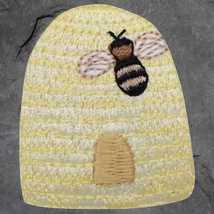 May include: Embroidered patch featuring a yellow beehive with a brown entrance and a black and brown bee with white wings. The patch has a textured appearance and is suitable for sewing or ironing onto fabric.