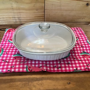 May include: A white ceramic baking dish with a glass lid sits on a pink and white checked hot pad with a watermelon print.
