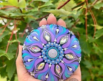 May include: A hand-painted, round stone featuring a mandala design. The mandala has a central silver circle surrounded by blue, purple, and teal petal-like shapes. Iridescent crystals are placed around the design. The stone is held against a backdrop of green leaves.
