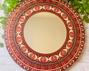 May include: Round decorative mirror with a detailed, hand-painted design. The border features a repeating floral pattern in shades of brown, red, and peach. The mirror's edge is adorned with small, evenly spaced dots. The mirror is a home decor item.