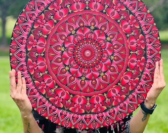 May include: A round, decorative mandala with a complex floral design. The mandala features a vibrant color palette of red, pink, and gold against a black background. The artwork is held up in front of a person.