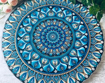 May include: A round, hand-painted mandala in shades of blue, teal, and gold on a black background. The intricate design features concentric circles with floral and dot patterns. The artwork is displayed against a backdrop of flowers and greenery.