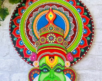 May include: A vibrant, hand-painted Kathakali mask. The mask features a green face with blue eyes, red lips, and a white mustache. The circular background is adorned with intricate patterns in red, green, blue, and yellow, creating a striking visual.