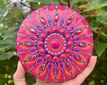 May include: A vibrant, hand-painted mandala stone with a detailed floral design. The stone features a central pink circle surrounded by concentric circles of pink, orange, and blue, embellished with red gems. The stone is held in a hand.