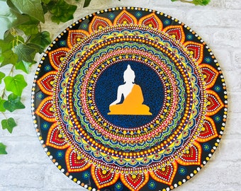 May include: A round, decorative wall art piece featuring a detailed mandala design. The artwork showcases a central image of a seated Buddha in orange and white, surrounded by concentric circles of colorful patterns. The background is a deep blue.