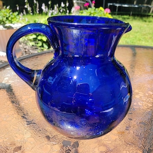 Puede incluir: Una jarra de vidrio azul cobalto con cuerpo redondeado y labio acampanado. La jarra tiene una sola asa y un borde dorado.