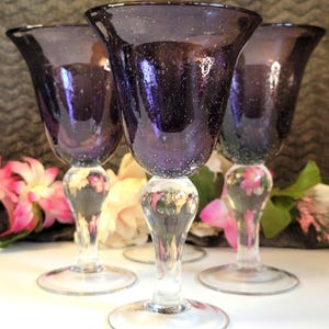 May include: Four elegant, purple-tinted wine glasses with clear, bulbous stems and round bases. The glass has a speckled texture. The glasses are arranged in a group, with flowers in the background, suggesting a formal setting.