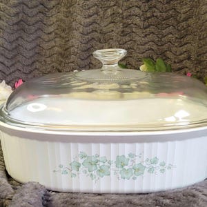 May include: White oval casserole dish with a clear glass lid. The dish has vertical ribbed sides and a floral design in light green. The lid has a clear glass handle.