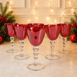 May include: A set of five wine glasses with deep red bowls and clear glass stems featuring a stacked ball design. Each glass has a wide, circular base and a smooth, rounded rim, arranged in a cluster on a light-coloured surface.