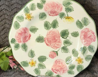May include: A decorative, off-white plate with a scalloped edge and a green border. The plate is adorned with pink roses, green leaves, and yellow flower accents. The plate is resting on a silver metal stand.
