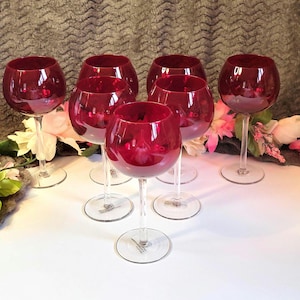 Pode incluir: Conjunto de seis taças de vinho tinto rubi. Cada taça apresenta uma taça arredondada e uma haste fina. As taças estão dispostas sobre uma superfície branca, com detalhes florais no fundo. As taças são de vidro.