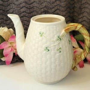 May include: Cream-colored ceramic teapot with a woven basket design. The handle is green and brown, and the spout is curved. Green shamrock accents decorate the body of the teapot. A charming piece for tea lovers.