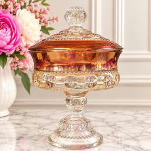 May include: A pedestal candy dish made of iridescent amber and clear glass. The bowl features a wide, rounded shape with a matching lid, decorated with a repeating circular pattern and a textured rim. The base is a sturdy, tiered glass stand.