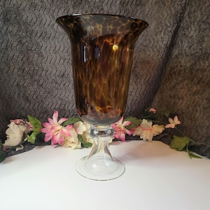 May include: A tall, elegant glass vase with a flared rim and a clear, pedestal base. The vase has a brown, speckled pattern throughout the body. The base is clear glass. Decorative flowers are arranged around the base of the vase.