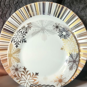 May include: White ceramic plates with gold and black striped rims. The smaller plate features snowflake designs in gold, silver, and black. A festive set for holiday dining or special occasions.