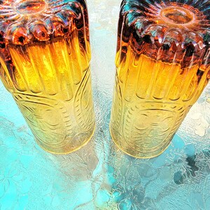 May include: Two amber glass tumblers with a textured, embossed design. The glasses have a yellow and orange gradient and a blue rim.