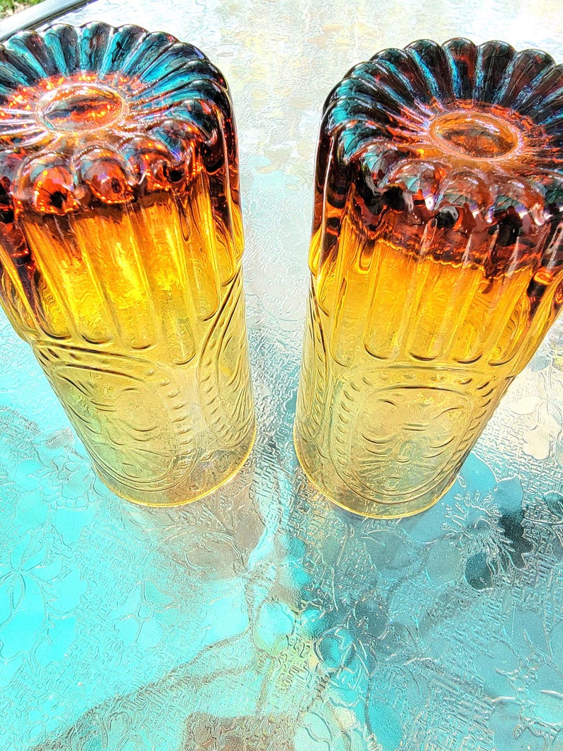 May include: Two amber glass tumblers with a textured, embossed design. The glasses have a yellow and orange gradient and a blue rim.