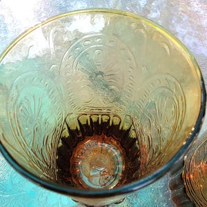 May include: A clear glass tumbler with an amber tint and an embossed floral design. The glass has a rounded bottom and a wide mouth.