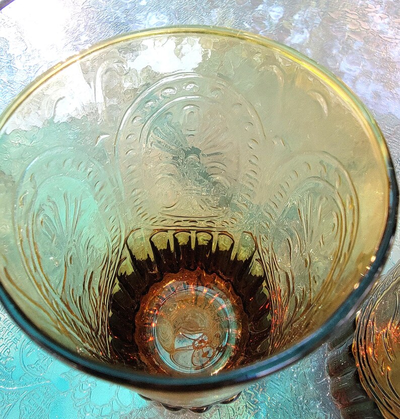 May include: A clear glass tumbler with an amber tint and an embossed floral design. The glass has a rounded bottom and a wide mouth.